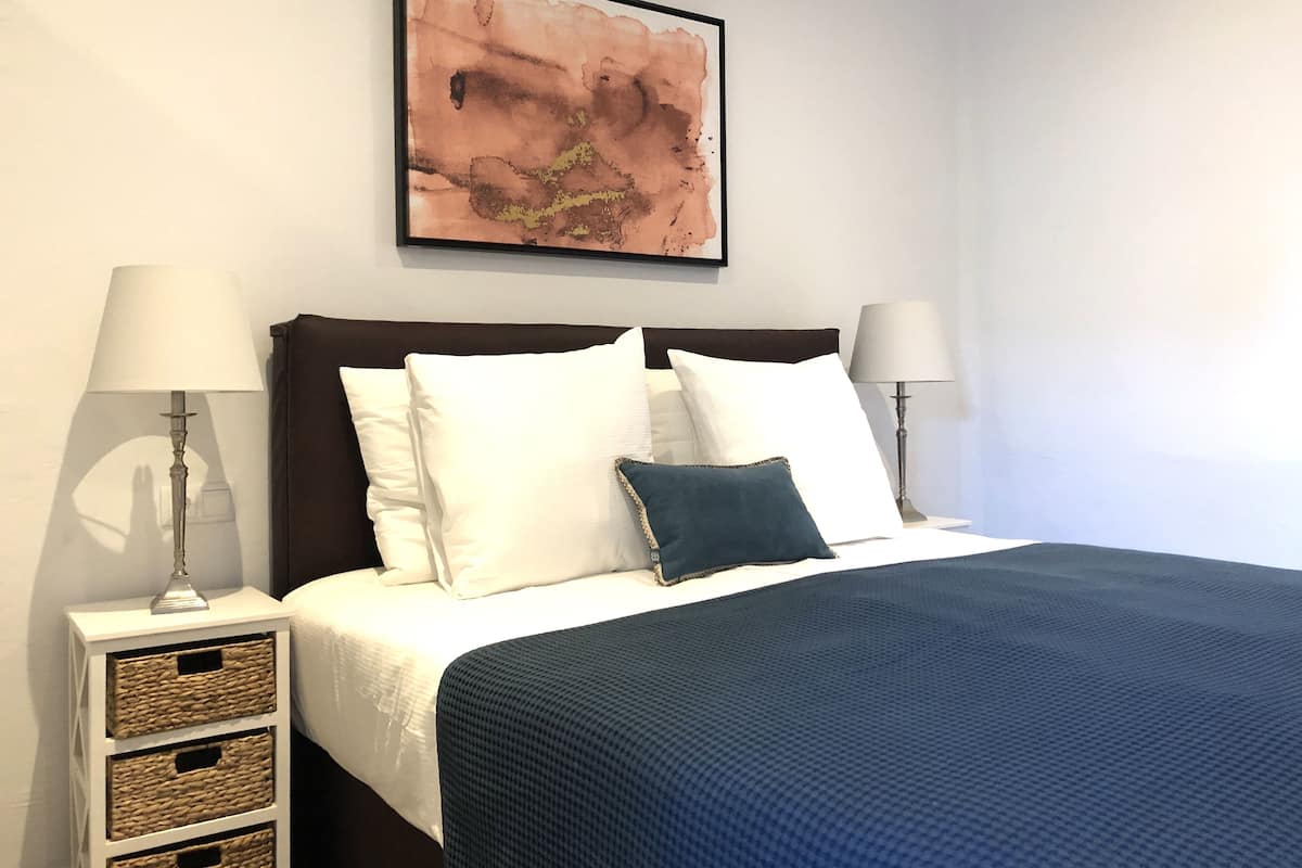A neatly made bed with white and blue bedding, two bedside lamps, and a framed abstract painting on the wall.