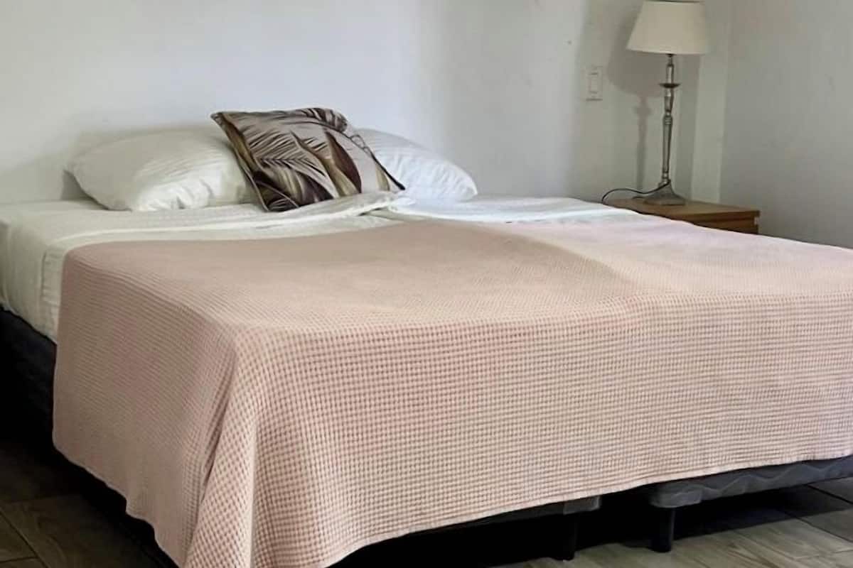 A room with a bed, a wall-mounted air conditioner, a lamp, and a floral painting.