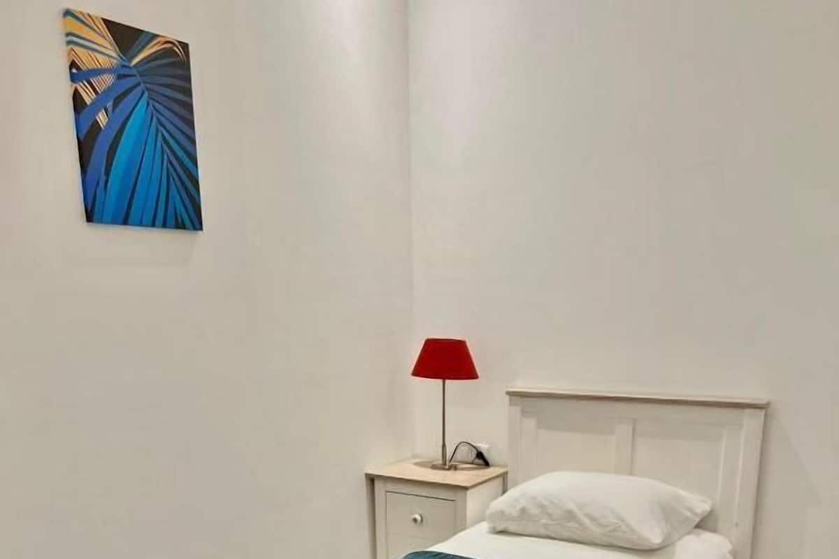 A neatly made bed with a white headboard and a teal bedspread, a small bedside table with a red lamp, and a wall-mounted abstract painting.