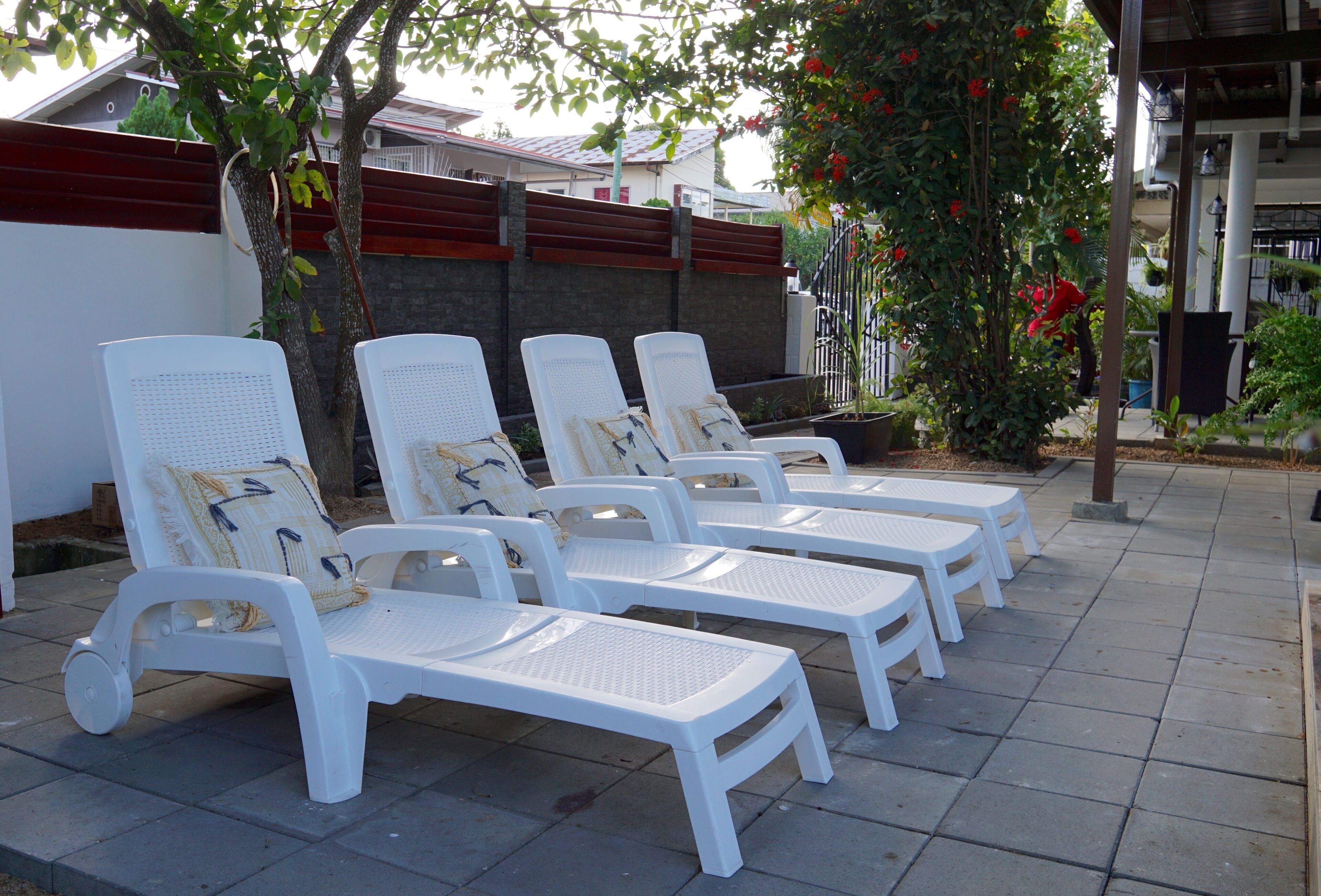 Een rij witte lounge stoelen met kussens, geplaatst op een geplaveid terras met een bakstenen muur en een boom met rode bloemen op de achtergrond.