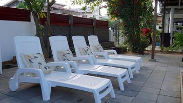 Een rij witte lounge stoelen met kussens, geplaatst op een geplaveid terras met een bakstenen muur en een boom met rode bloemen op de achtergrond.