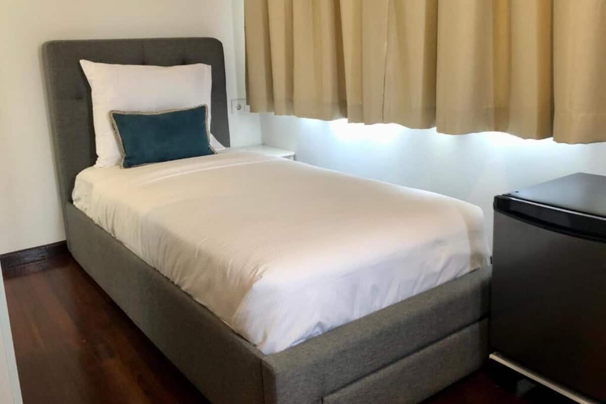 A single bed with a grey headboard, white bedding, and a teal accent pillow. A small black desk is to the right.