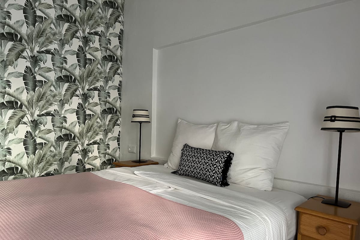 A neatly made bed with a pink blanket, white pillows, and a black and white patterned pillow. There are two bedside tables with lamps, a wooden nightstand, and a wall with a leaf-patterned wallpaper.