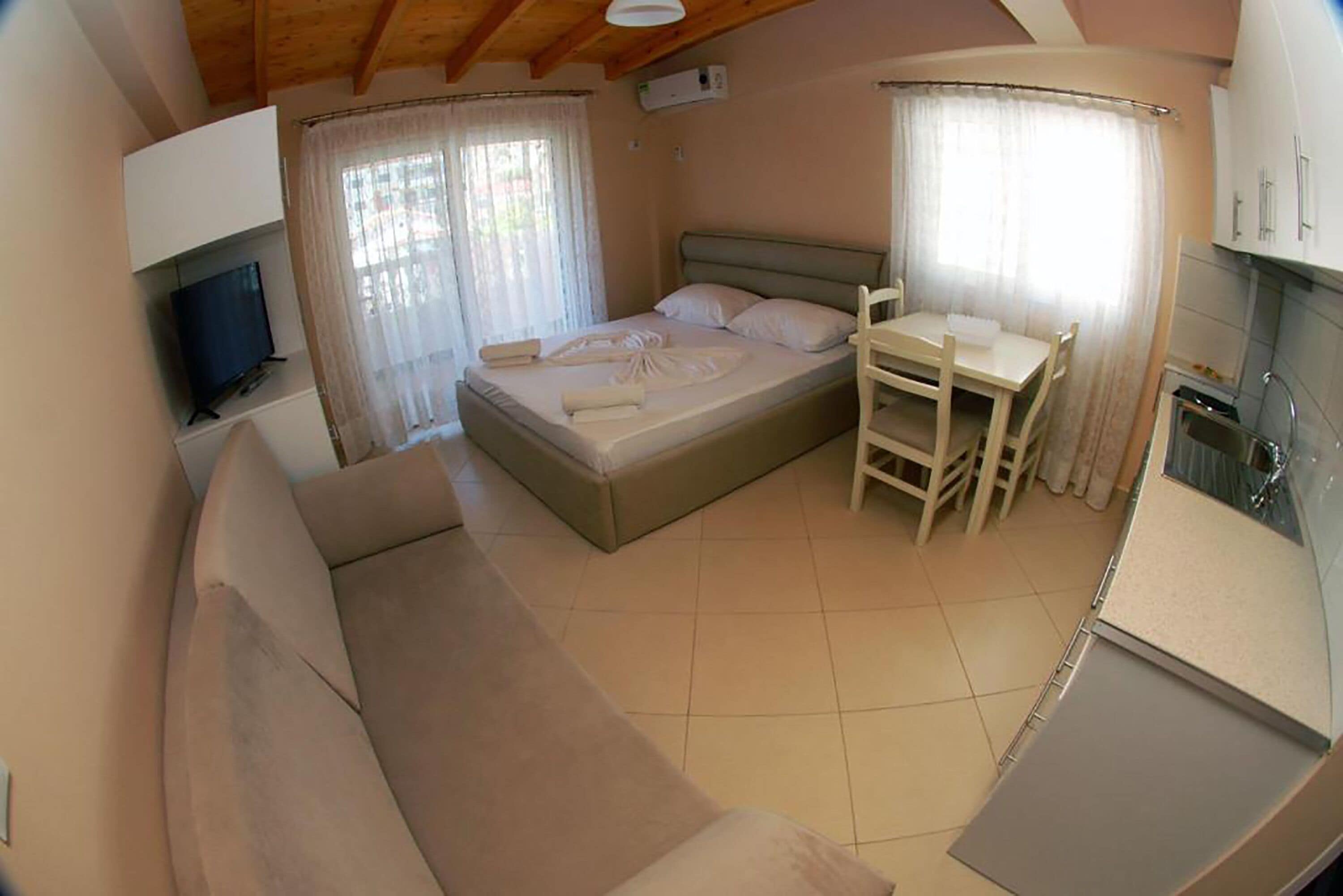 A bedroom with a bed, a small table, a chair, a TV, and a kitchenette.