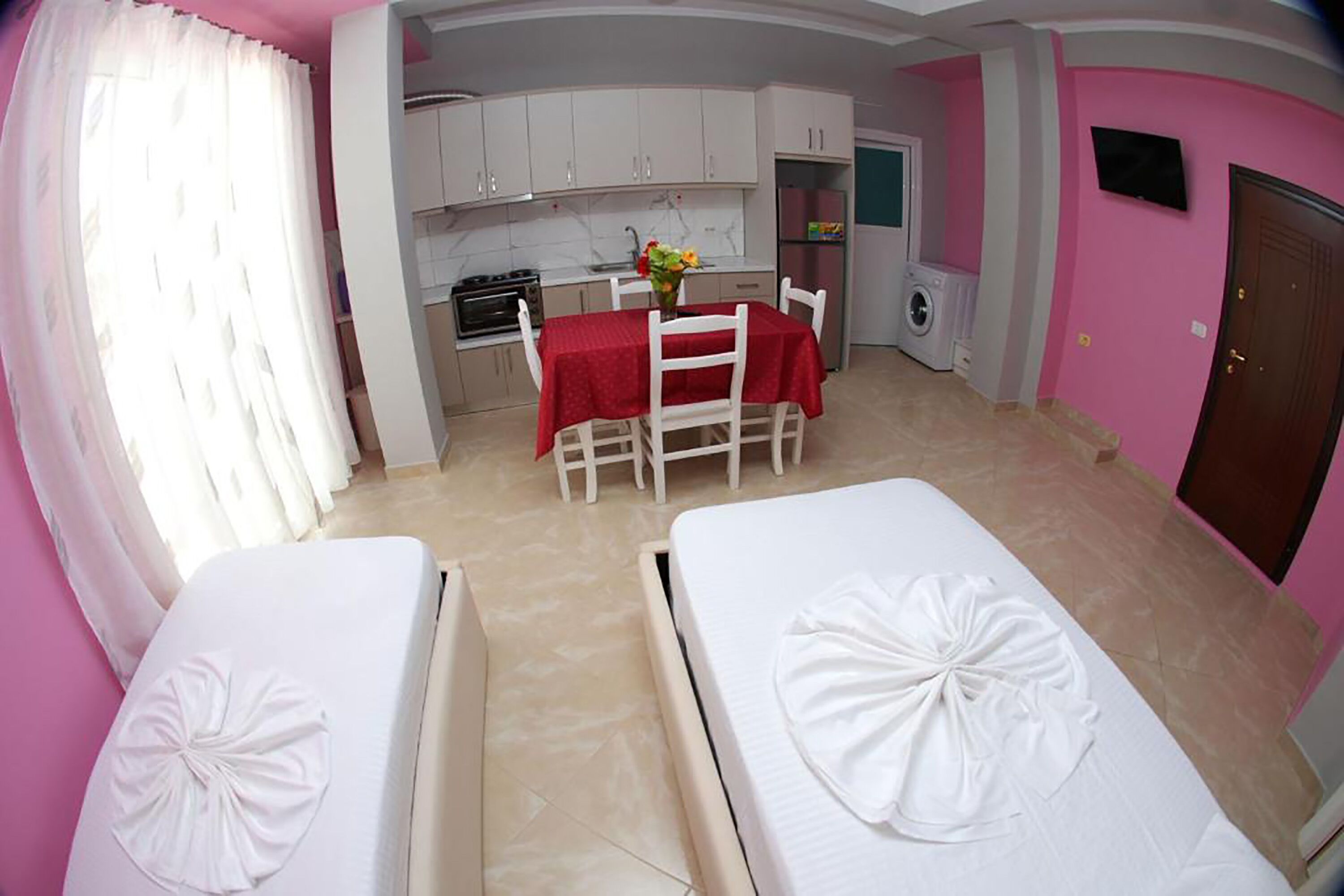 A room with two beds, a dining table with a red tablecloth, a kitchen area, and a pink wall.