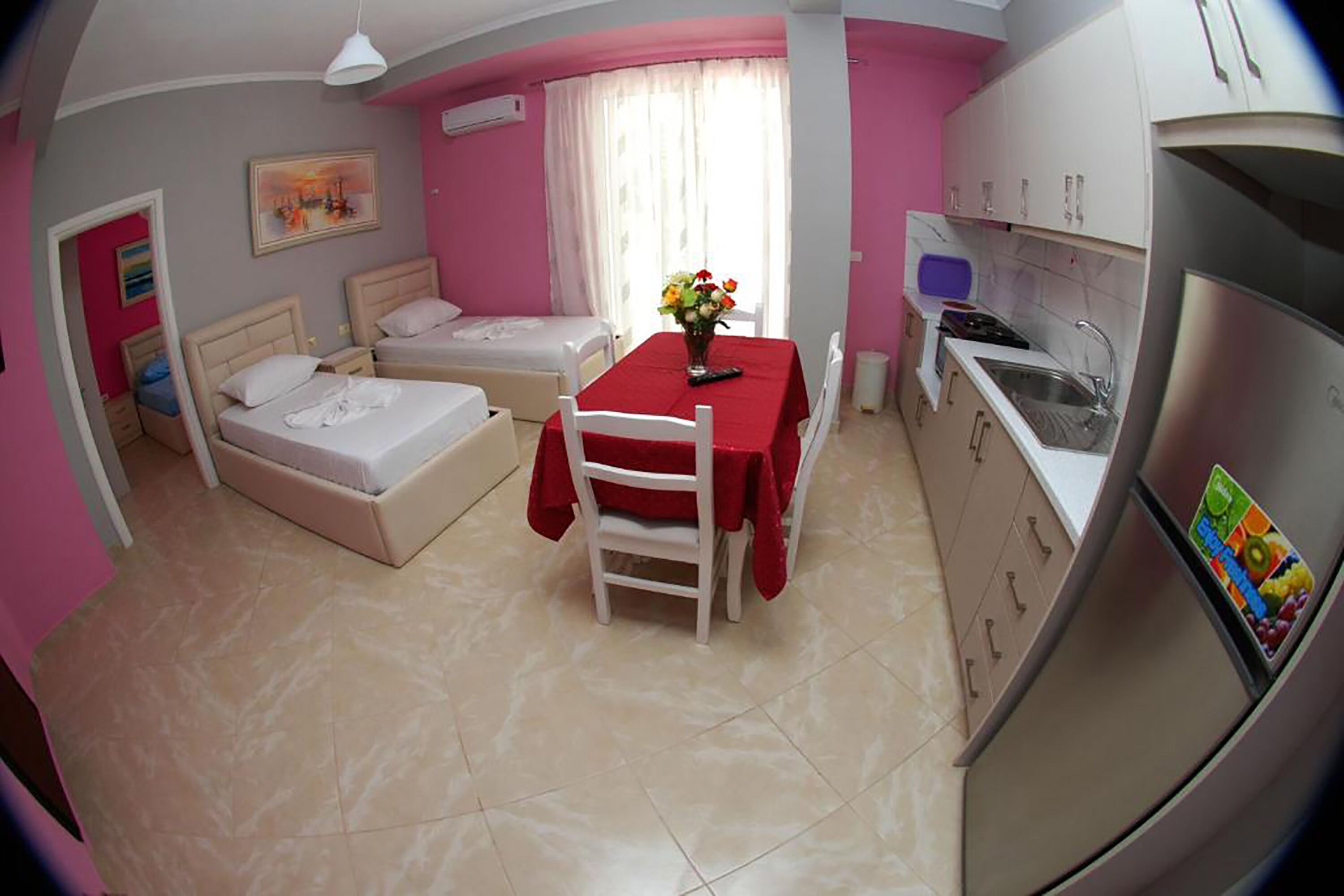 Una habitación compacta con cama, una mesita con un florero, kitchenette y ventana con cortinas.