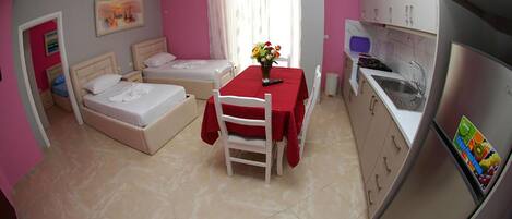 Una habitación compacta con cama, una mesita con un florero, kitchenette y ventana con cortinas.