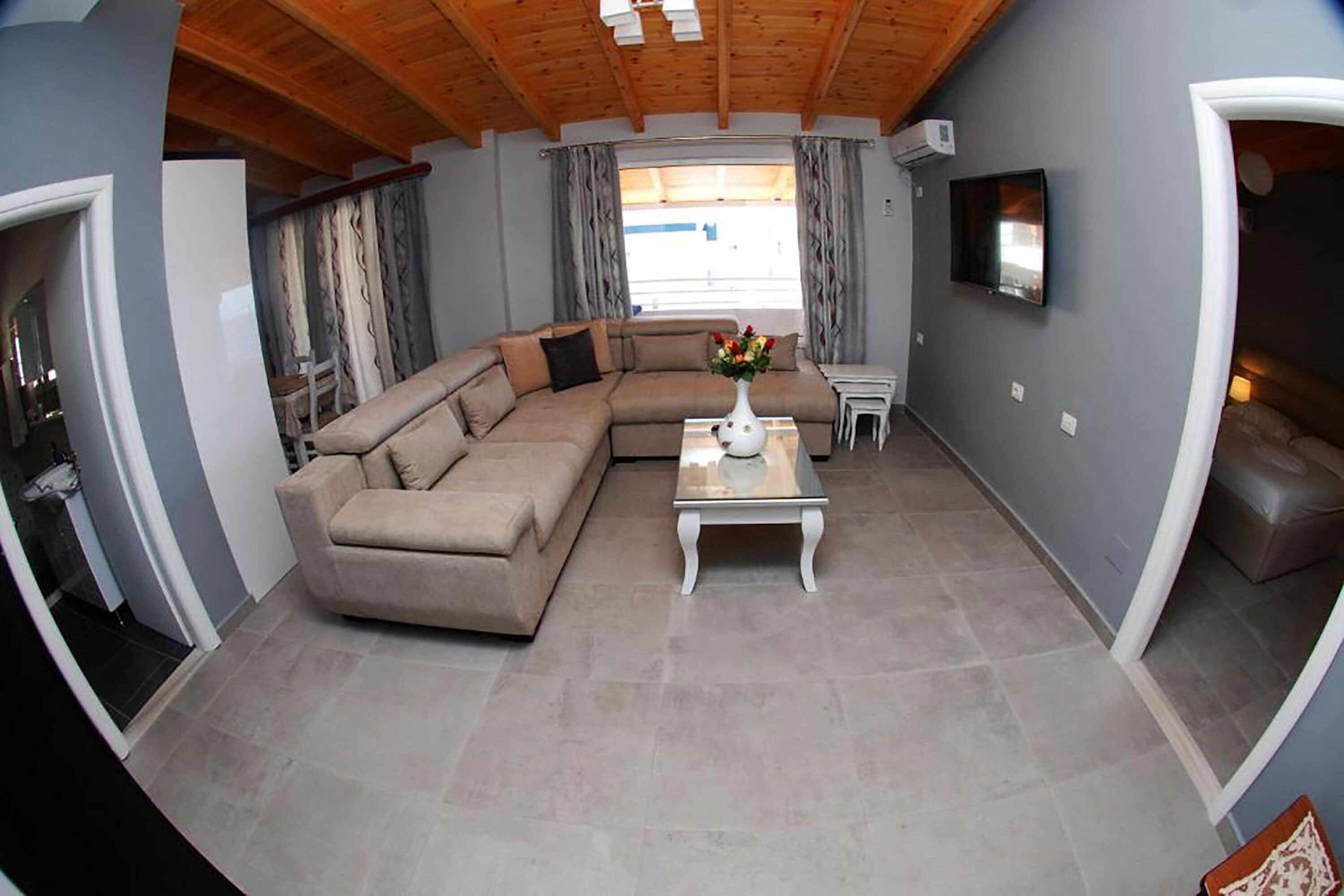 A living room with a beige sectional sofa, a white coffee table with a vase, and a flat-screen TV on the wall.