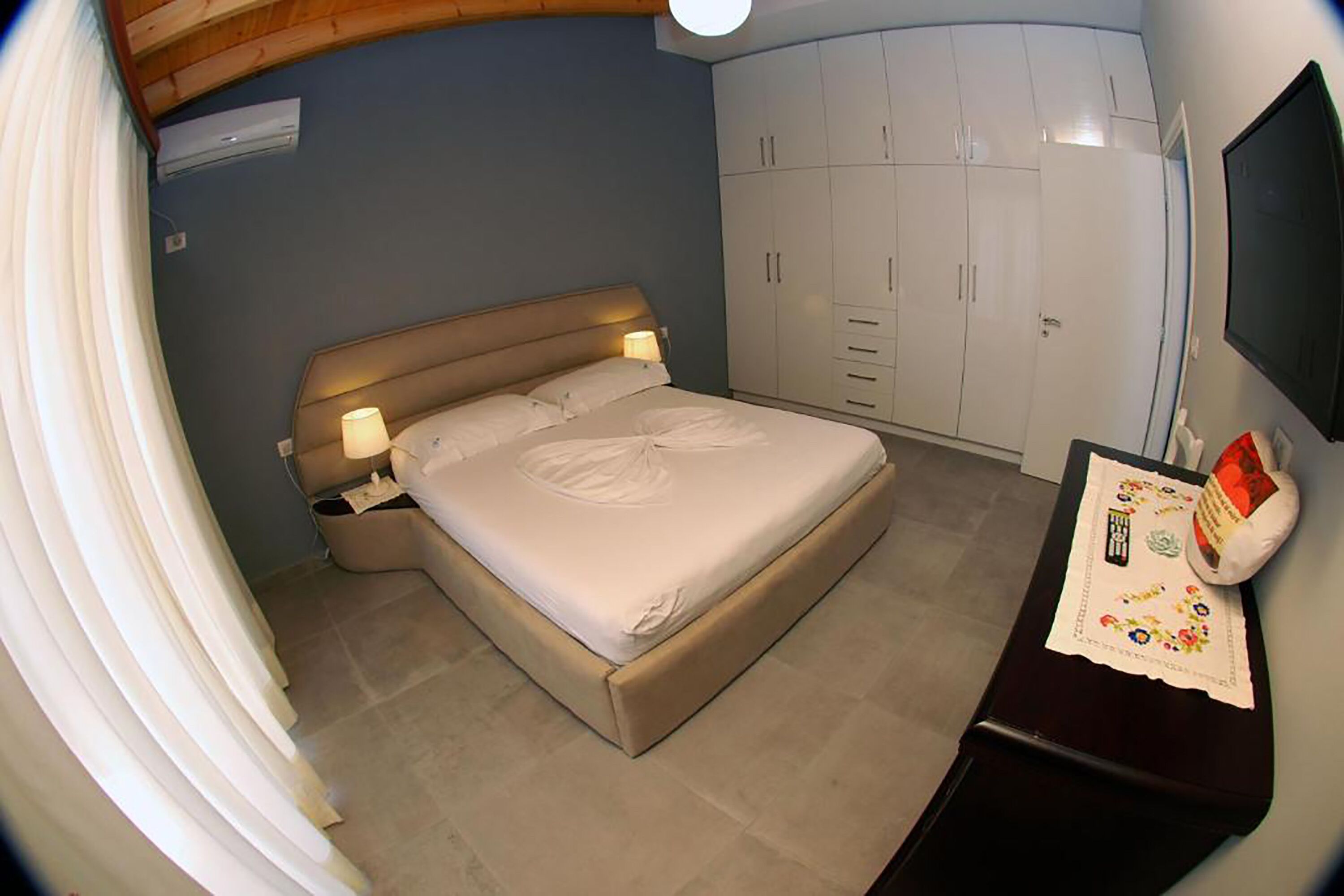 A bedroom with a bed, bedside lamps, a wardrobe, a desk with a lamp, and a curtain.