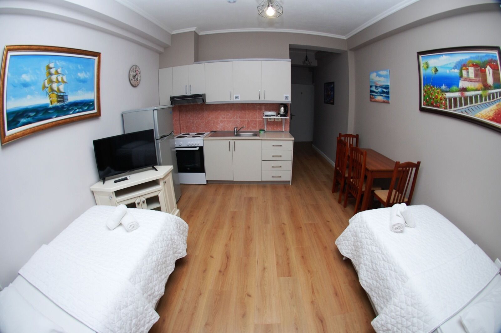 A compact hotel room with two beds, a kitchenette, a dining area, and artwork on the walls.