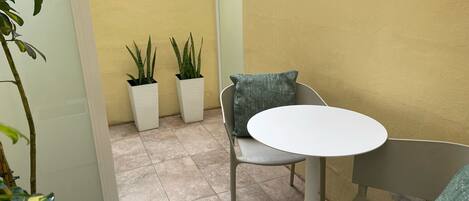 A small outdoor seating area with a round table and two chairs, two potted plants, and a yellow wall in the background.