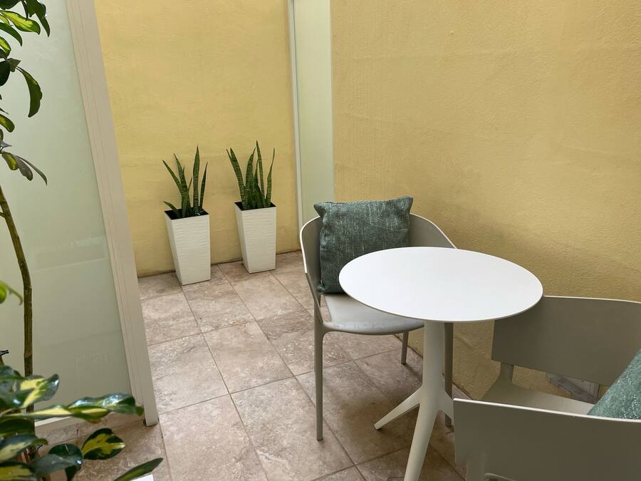 Ein kleiner Sitzbereich im Freien mit einem runden Tisch, zwei Stühlen, zwei Kübelpflanzen und einer gelben Wand im Hintergrund.