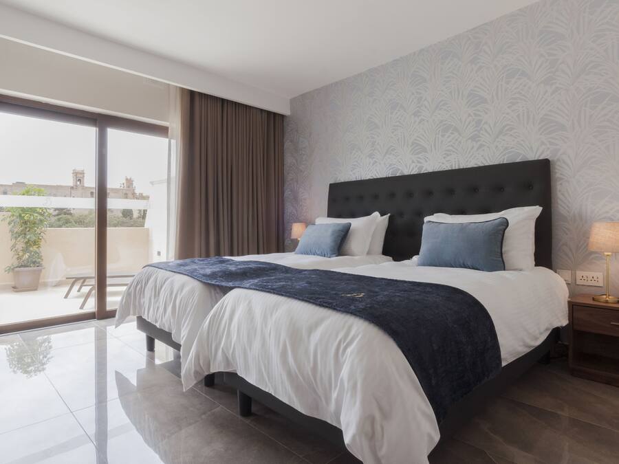 A modern hotel room with a large bed, a nightstand, and a view through a sliding glass door to an outdoor area.