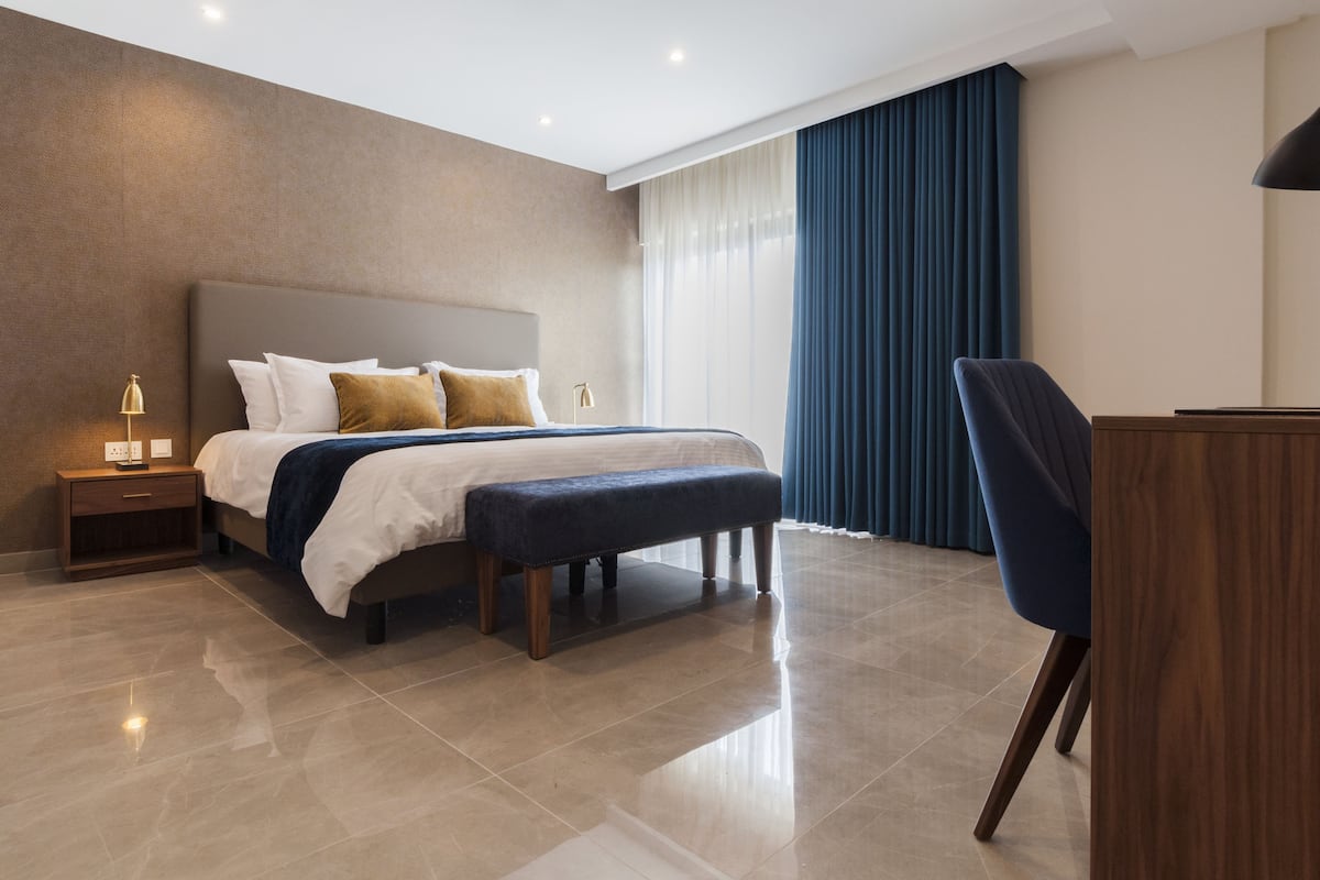 A modern hotel bedroom with a large bed, a bench, a desk, and a nightstand.