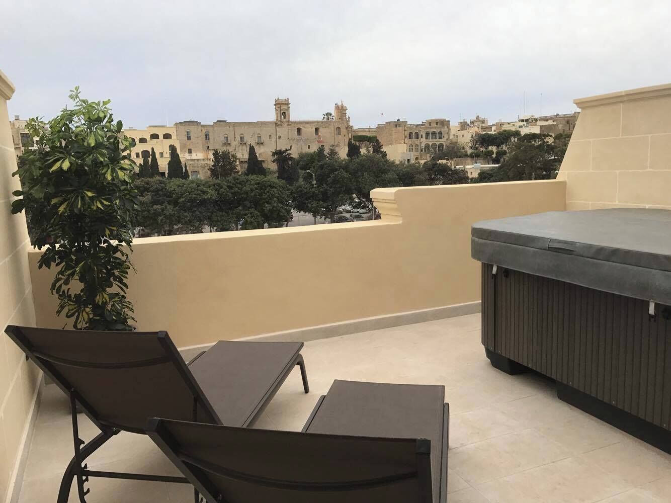 A rooftop terrace with two lounge chairs, a small table, and a hot tub. The terrace overlooks a cityscape with historic buildings.