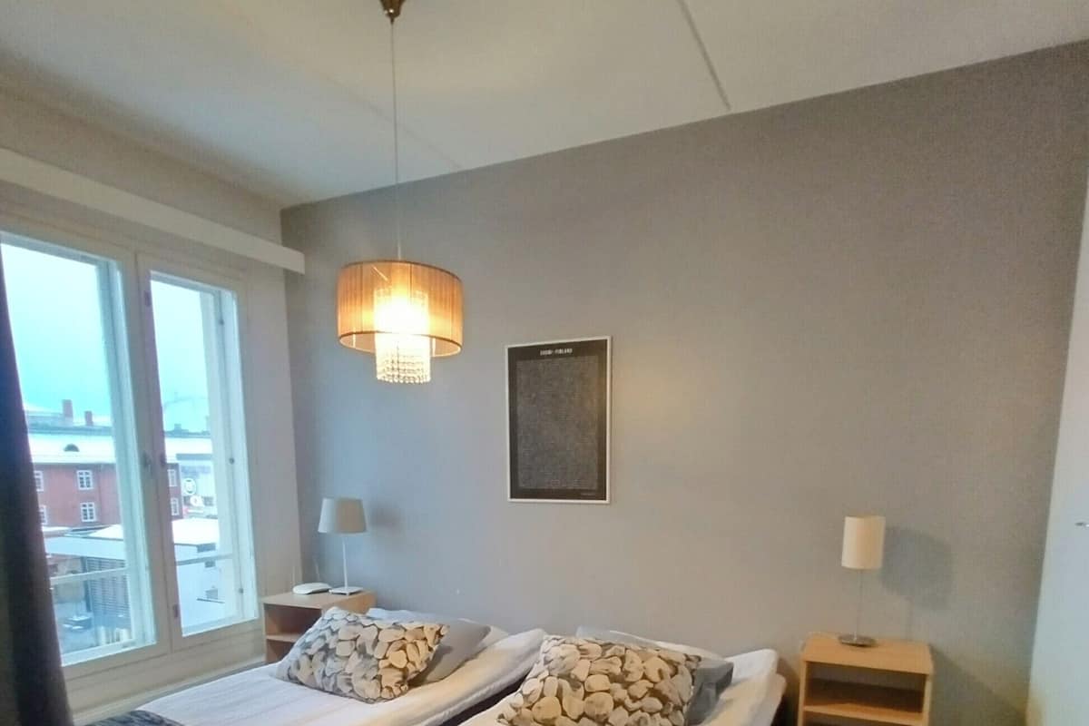 Una habitació amb dos llits, una petita taula de nit de fusta, una làmpada penjada a la paret i una finestra amb vista a edificis.