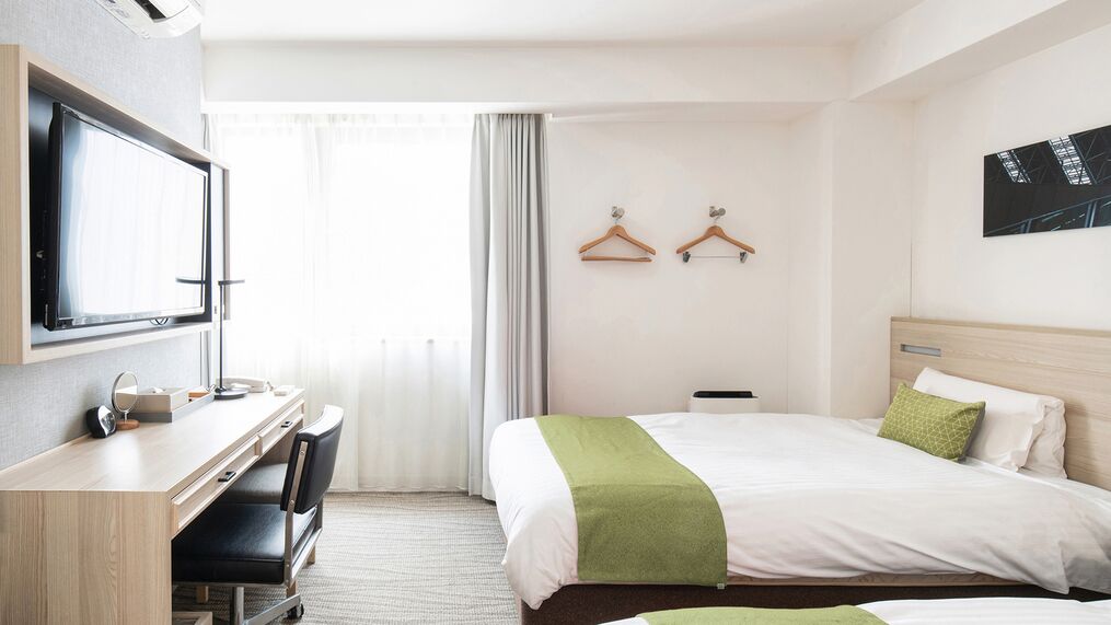 A hotel room with a bed, a desk with a chair, a television, and a window with curtains.