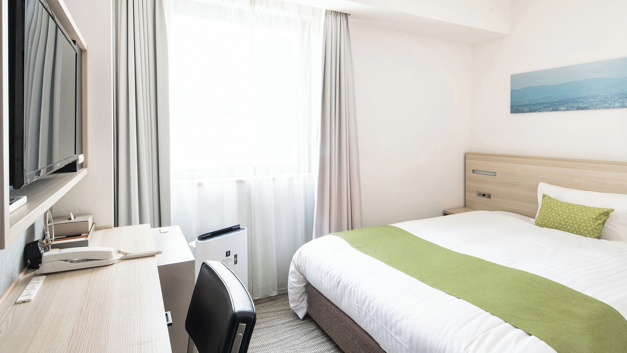 A hotel room with a bed, a desk with a computer, a TV, and a window with curtains.