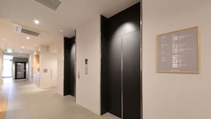 Corredor com elevador moderno, painel informativo fixado na parede e porta que dá para outro quarto.