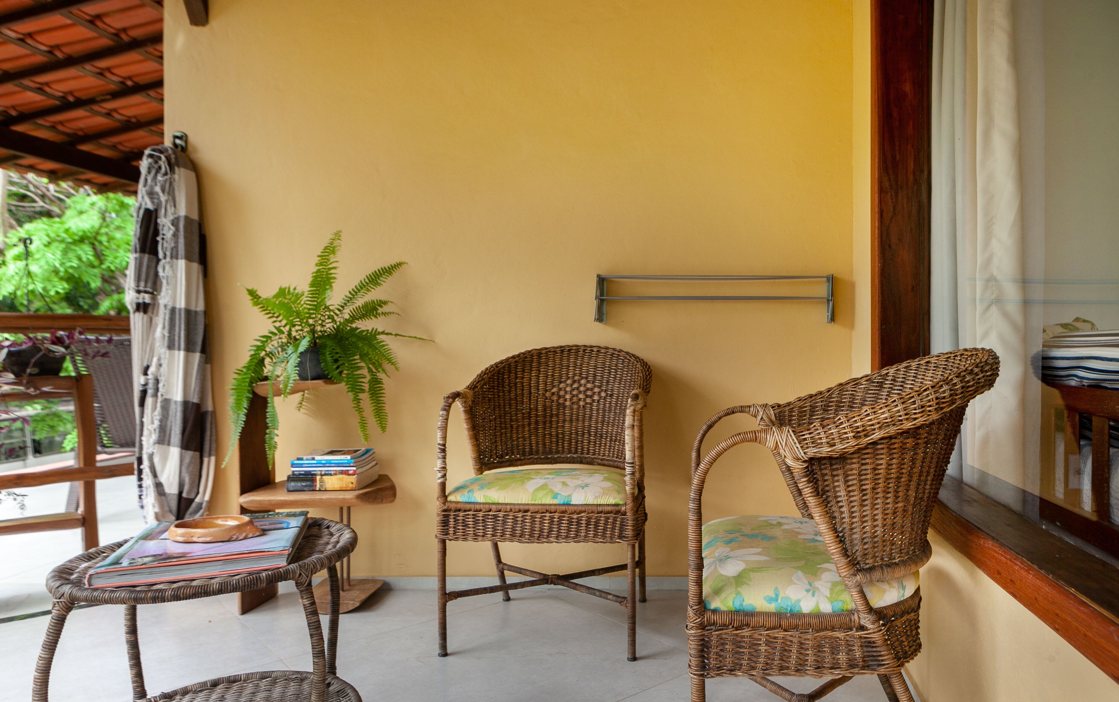A wicker chair with a floral cushion, a small round table with books and a bowl, and a potted fern plant.