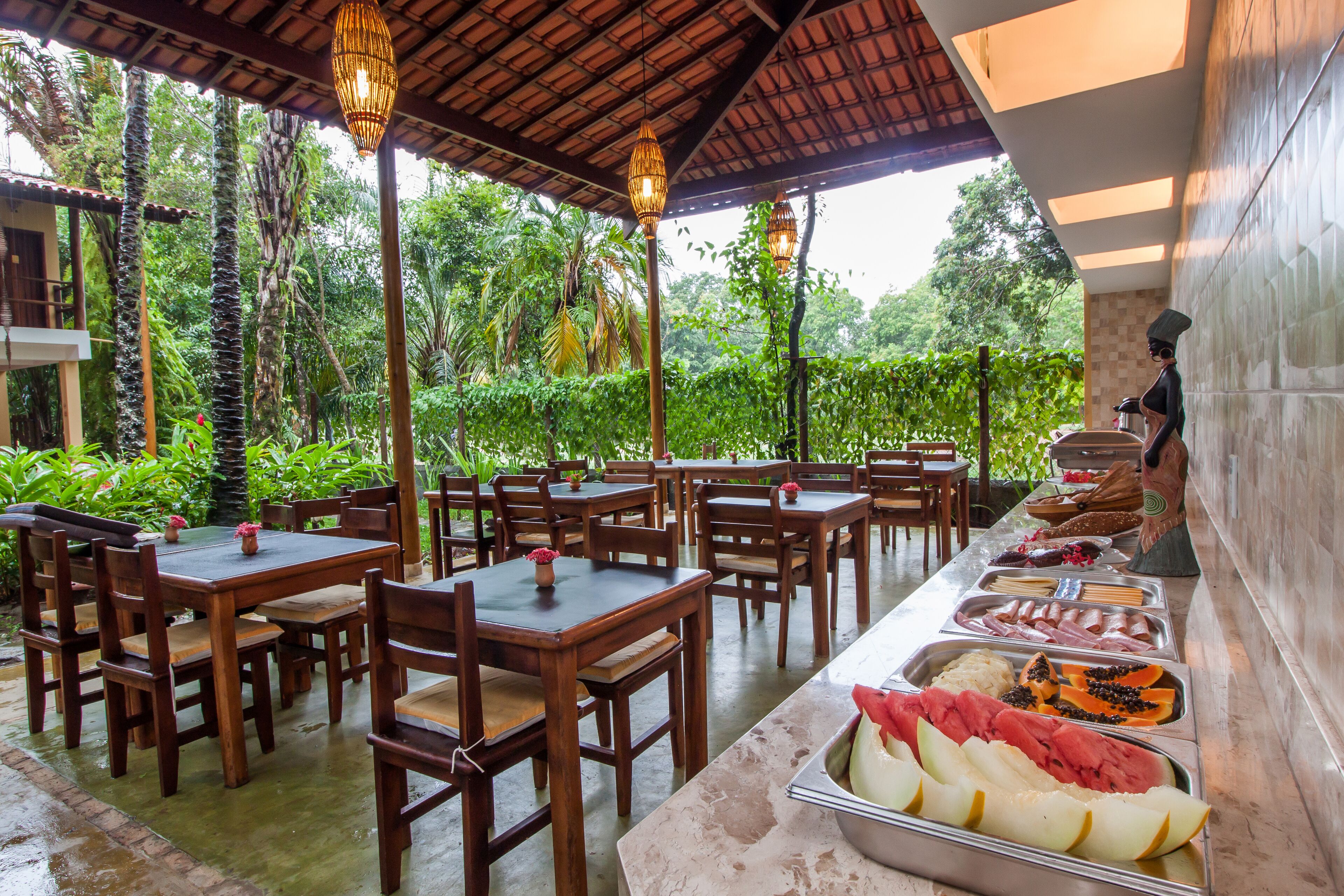 An outdoor dining area with wooden tables and chairs, surrounded by lush greenery.