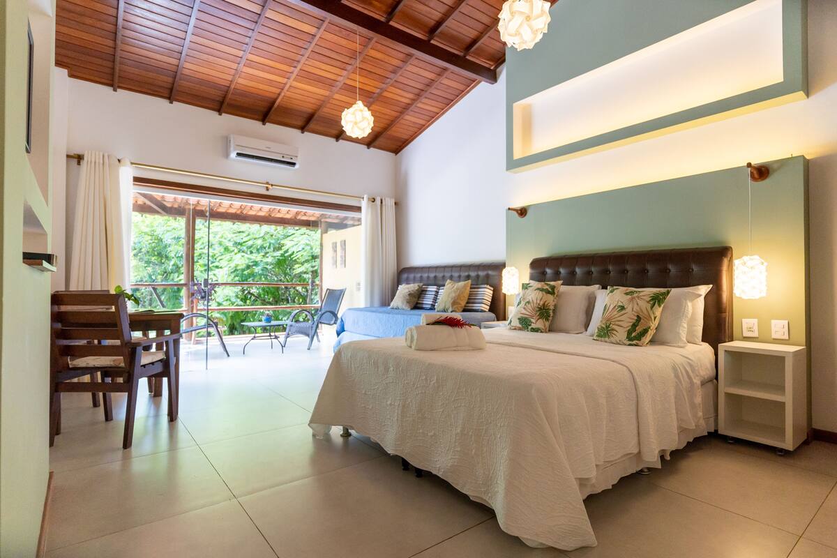 A spacious bedroom with a large bed, a wooden ceiling, and a view of greenery outside.