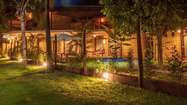 Un espacio exterior bien iluminado con piscina, palmeras y una zona de estar cubierta.