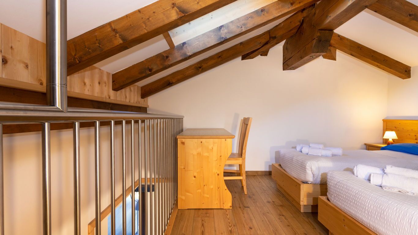 A cozy attic bedroom with a sloped ceiling, wooden beams, a small desk, a chair, and two beds.