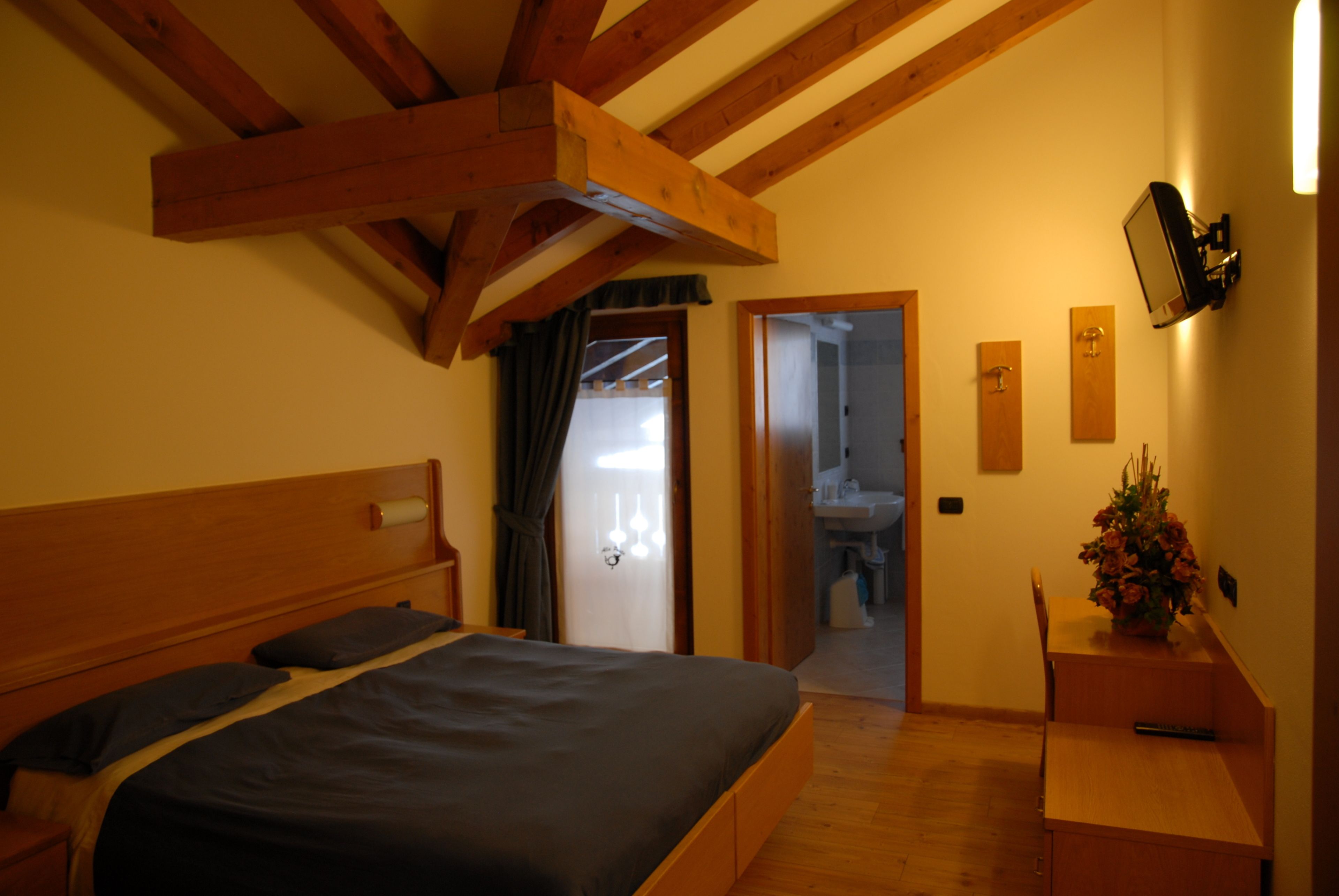 Een slaapkamer met een houten plafond, een bed met donker dekbed, een houten nachtkastje, een bijzettafeltje, een klein houten bureau met een bloevas en een tv aan de muur.