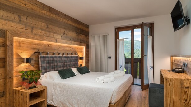 A bedroom with a wooden headboard, a bed with white linens, a bedside table with a lamp, and a view through a glass door to an outdoor area. - Albergo Alla Posta (Pinzolo)