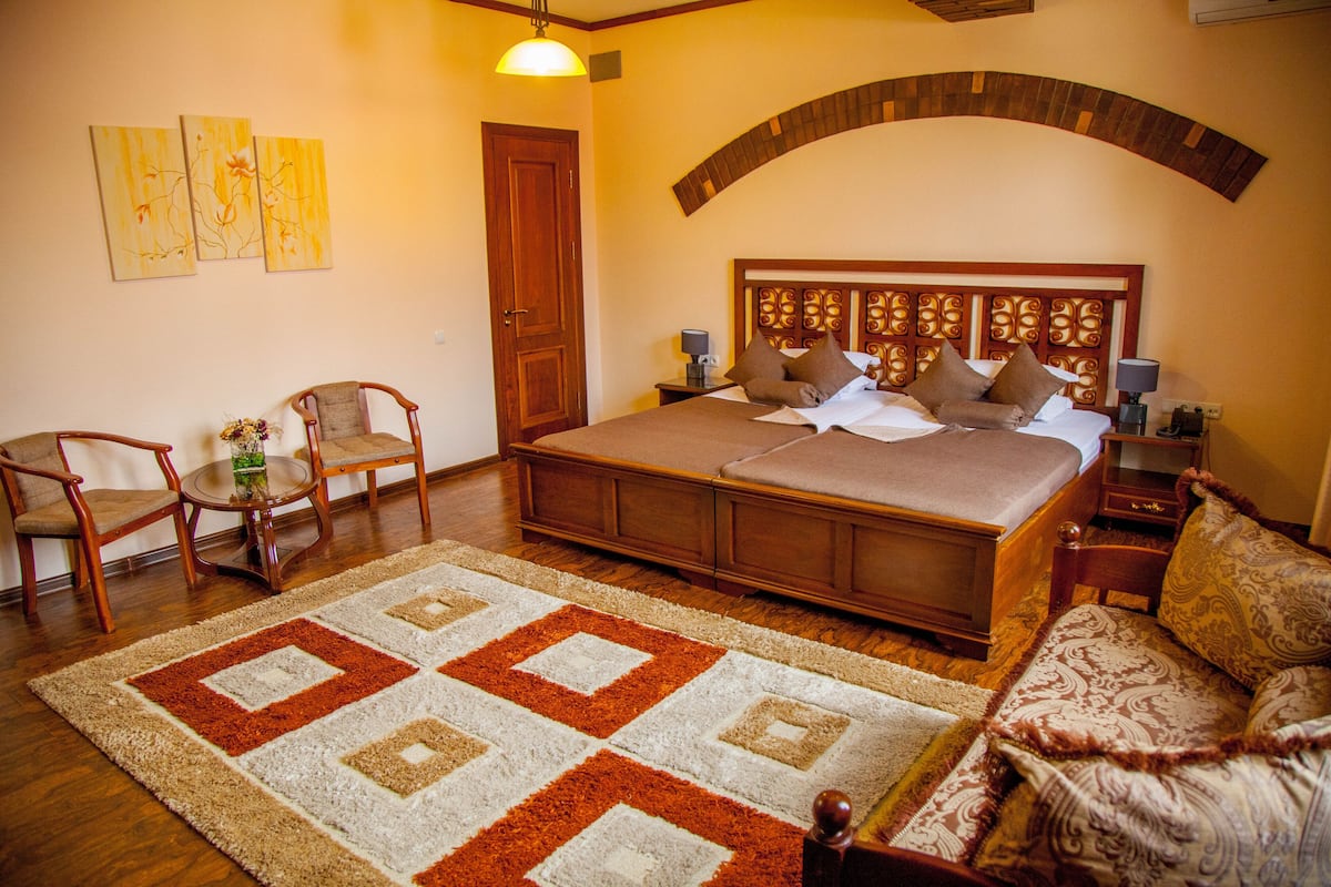 A bedroom with a large bed, a wooden headboard, a nightstand, two chairs, a rug, and artwork on the wall.