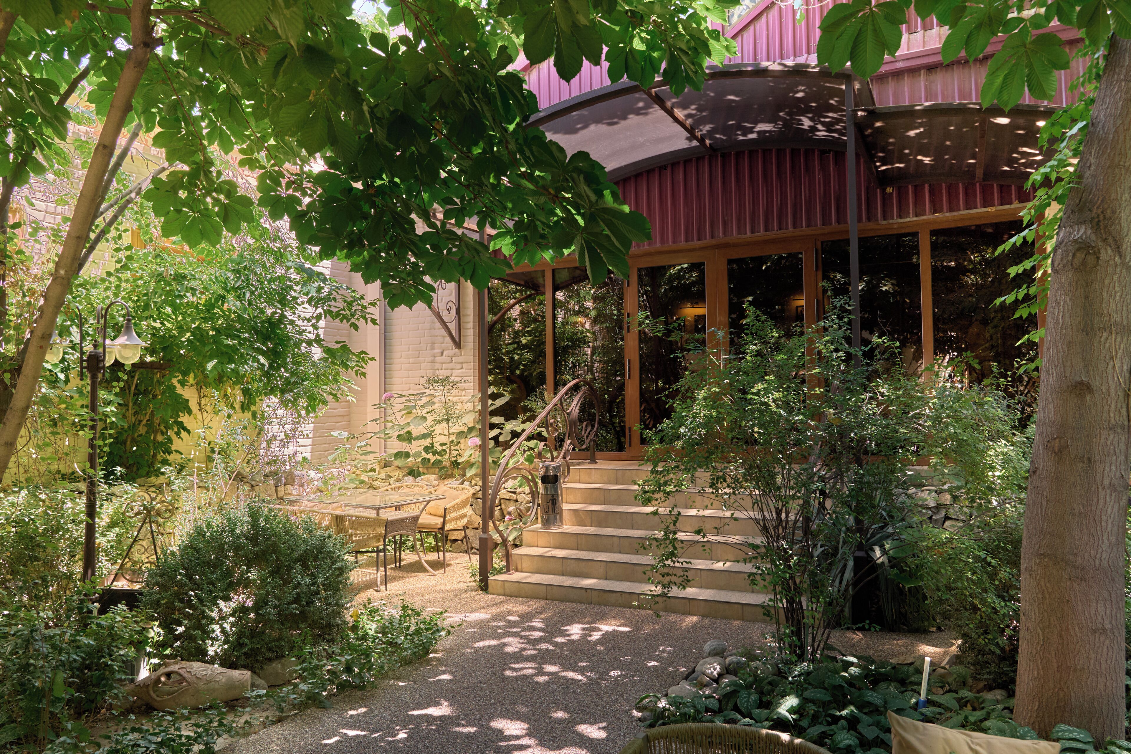 A covered outdoor seating area with stone steps leading up to a wooden door, surrounded by lush greenery and a stone wall.