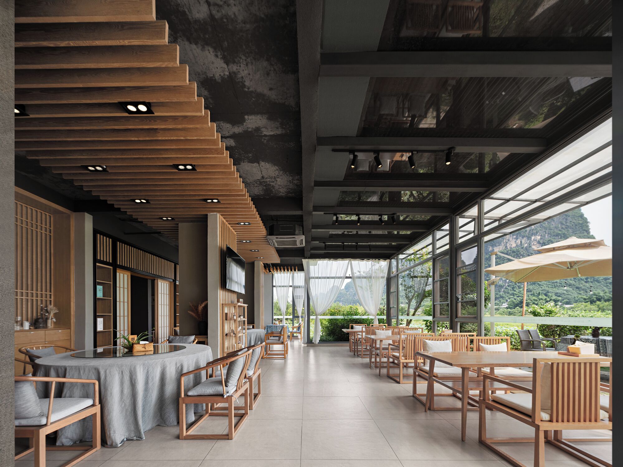 A modern restaurant with wooden furniture, large windows, and a view of greenery.
