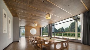 A spacious room with a large wooden table and chairs, a round table with a vase, and large windows offering a view of mountains.