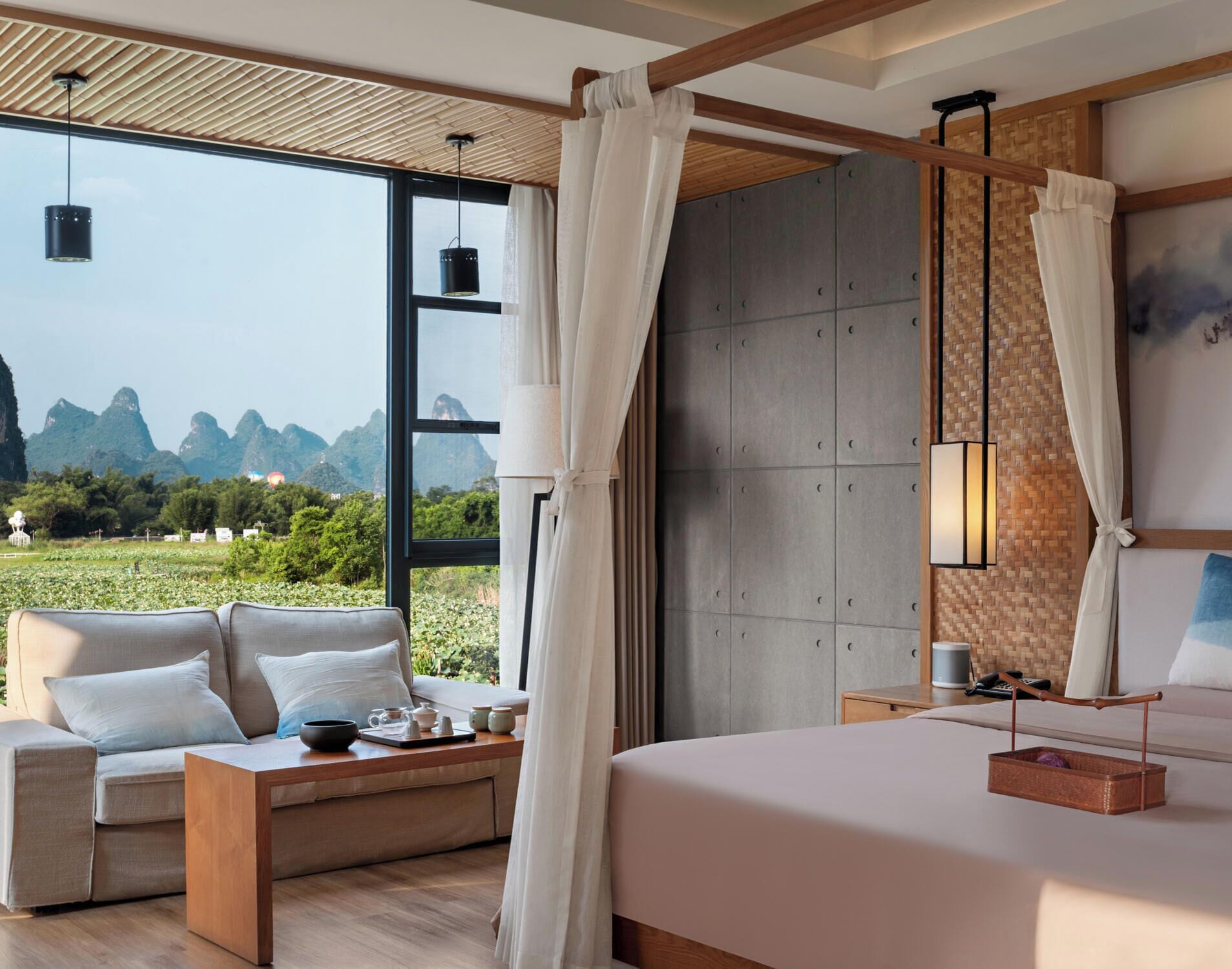 A modern hotel room with a large window offering a view of the mountains. There is a bed, a sofa, and a small table with tea set.
