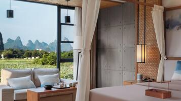 A modern hotel room with a large window offering a view of the mountains. There is a bed, a sofa, and a small table with tea set.