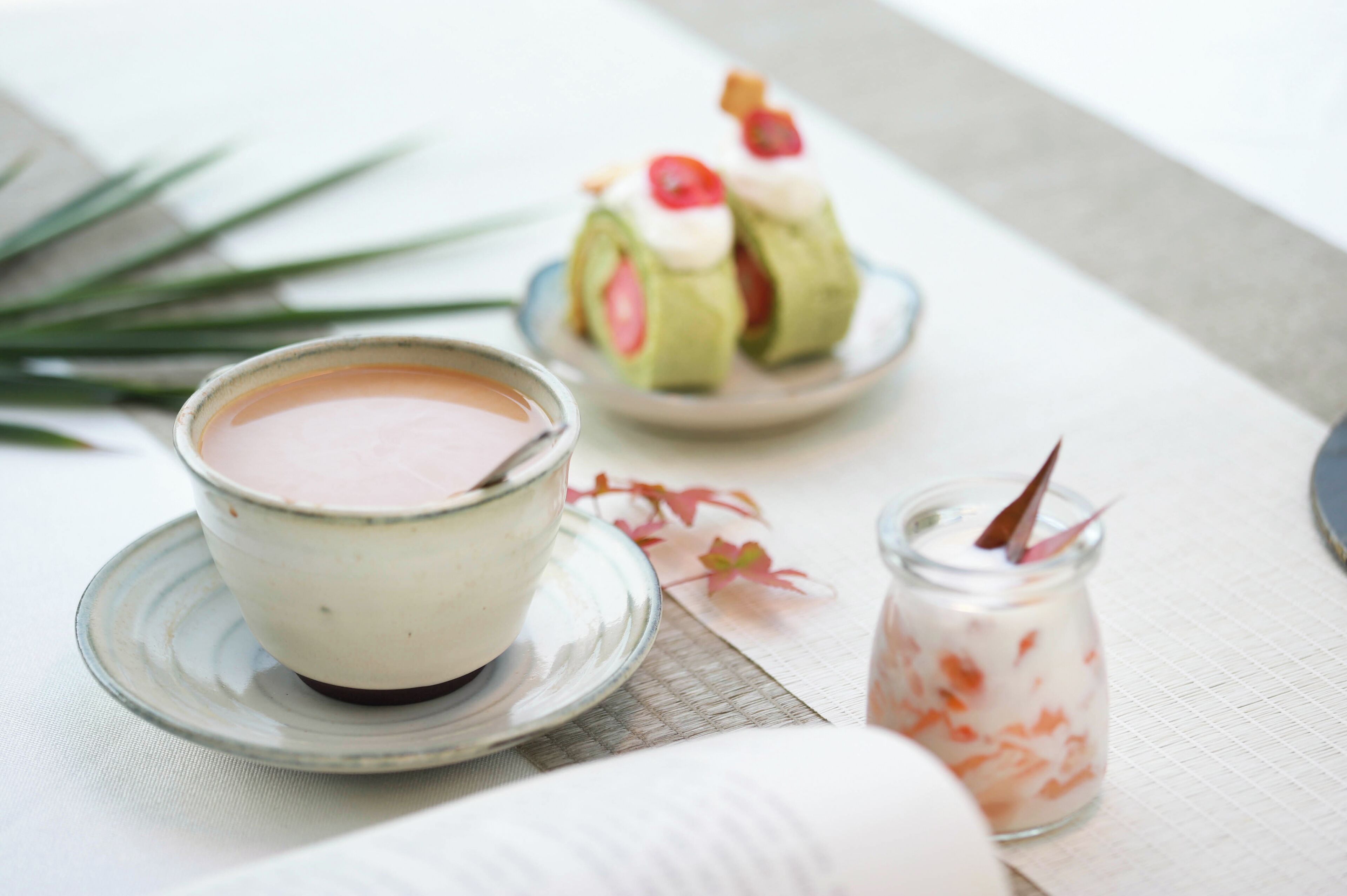 Ein Tisch mit einer Tasse Tee, einem Teller mit einem verpackten Dessert und einem kleinen Glas einer cremigen Substanz.