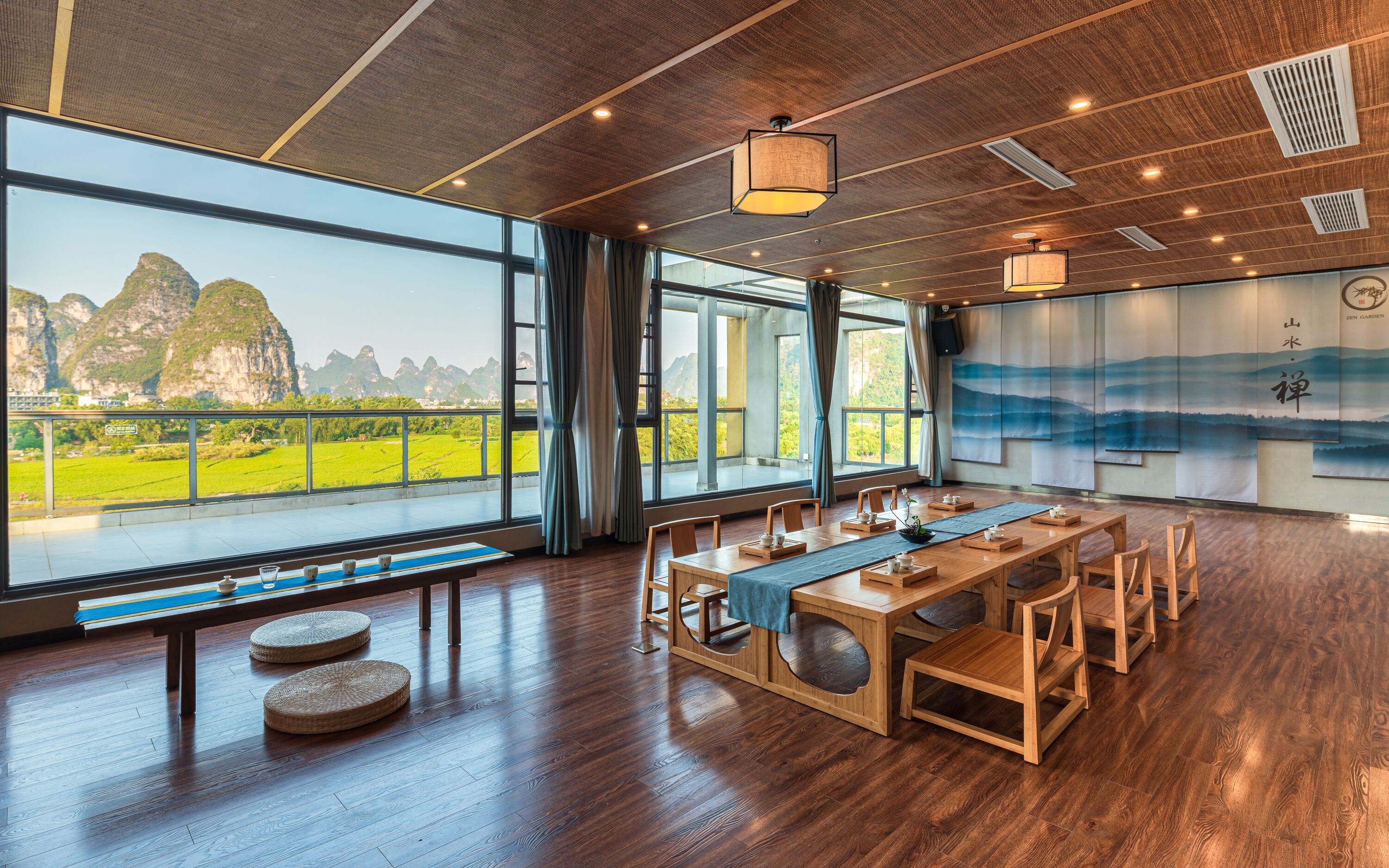 A spacious room with a long wooden table, multiple chairs, and a large window offering a view of green fields and mountains.