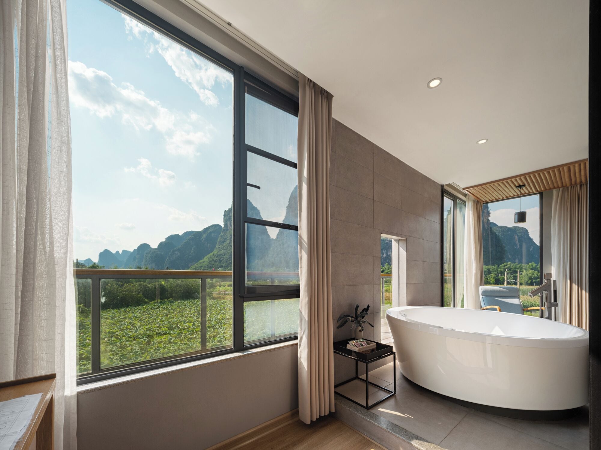 A modern bathroom with a large bathtub, a window with a view of mountains, and a small side table with a plant.
