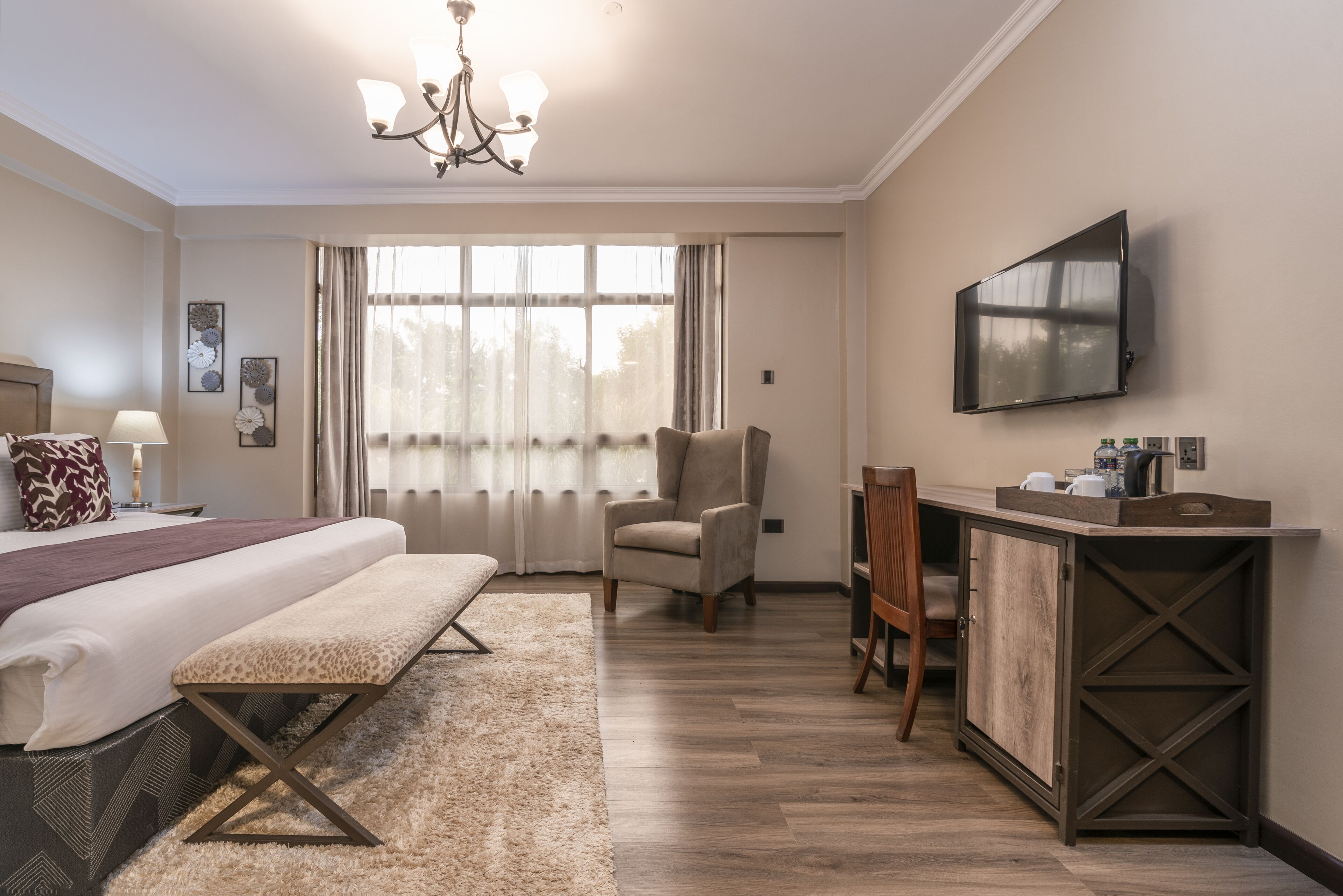 Habitación de hotel con una cama grande, un banco, una silla, un escritorio con televisor y una ventana con cortinas.