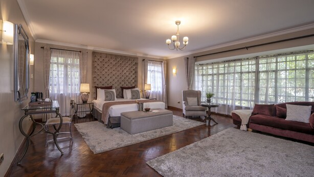 A spacious bedroom with a large bed, a sitting area with a sofa and armchair, a desk with a chair, and a large window providing natural light. - Fair Acres Boutique Hotel (Nairobi)