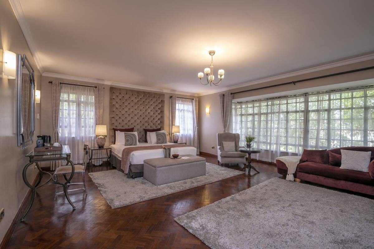 A spacious bedroom with a large bed, a sitting area with a sofa and armchair, a desk with a chair, and a large window providing natural light.