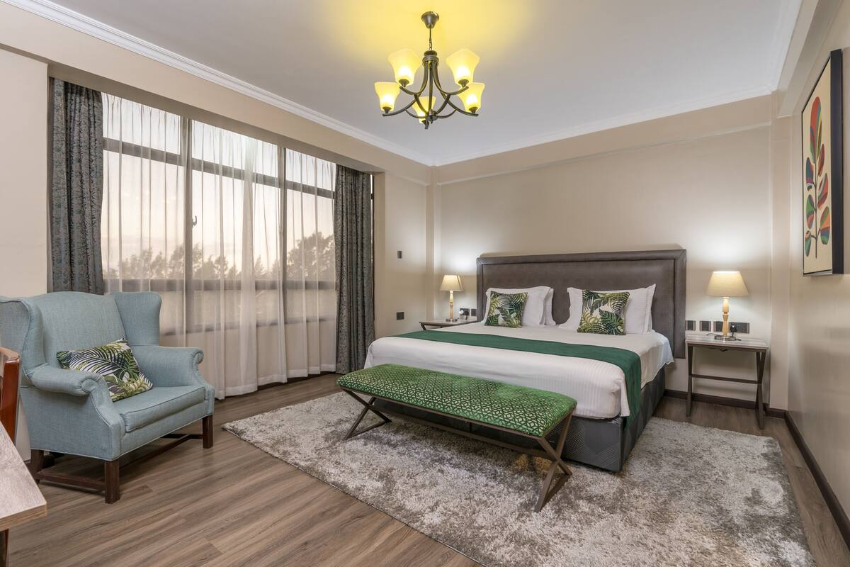 A hotel room with a large bed, a bench, a chair, a nightstand, and a chandelier.
