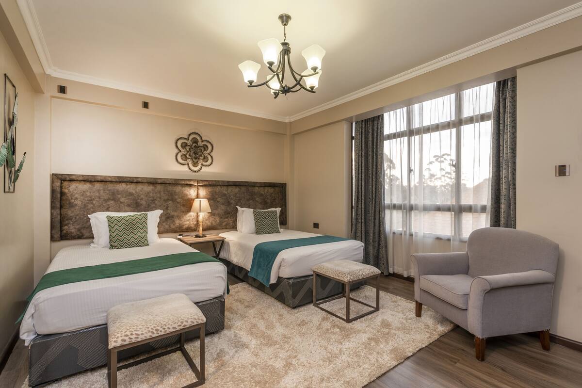 A hotel room with two beds, a chandelier, a large window with curtains, and a grey armchair.