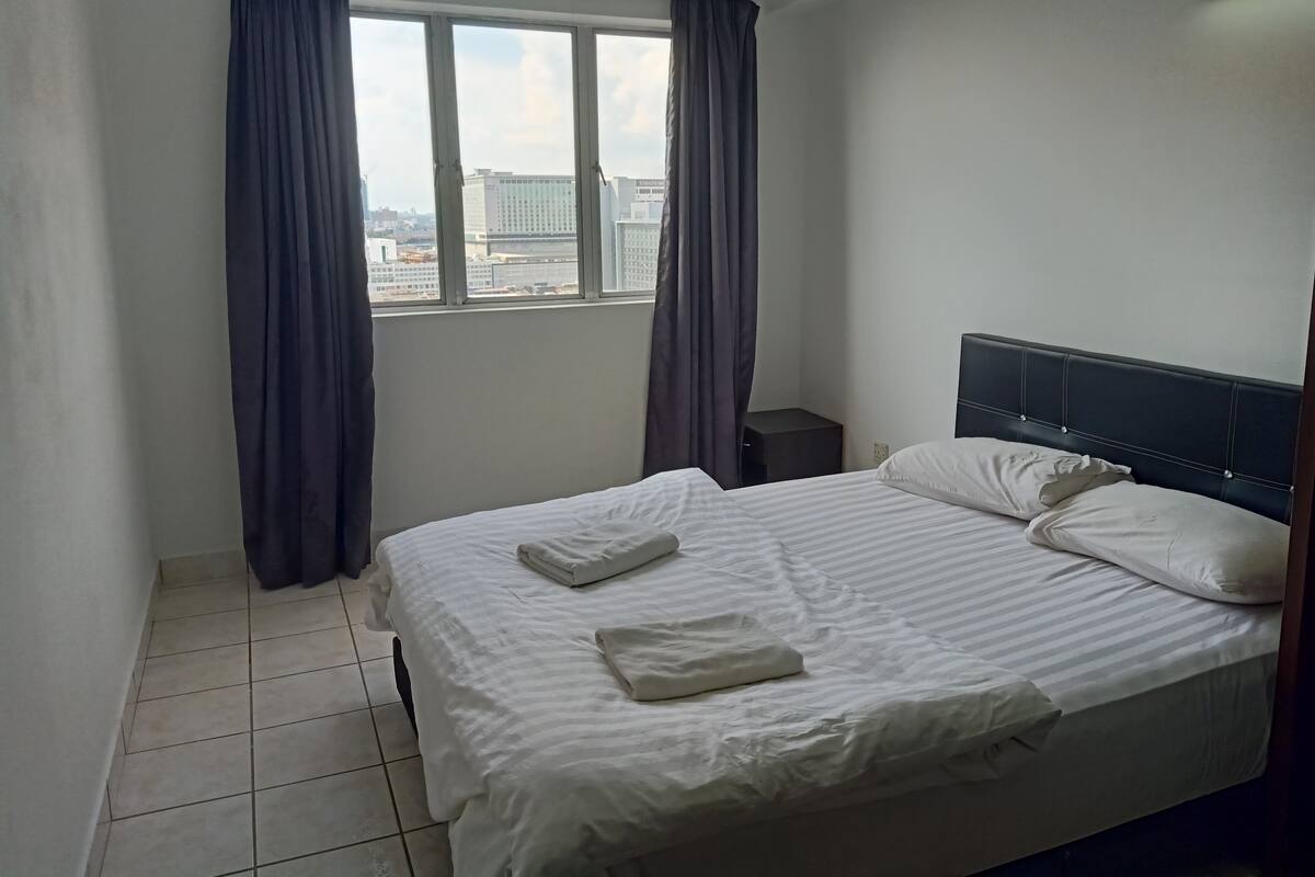 A hotel room with a bed, a nightstand, a window with curtains, and a view of buildings outside.