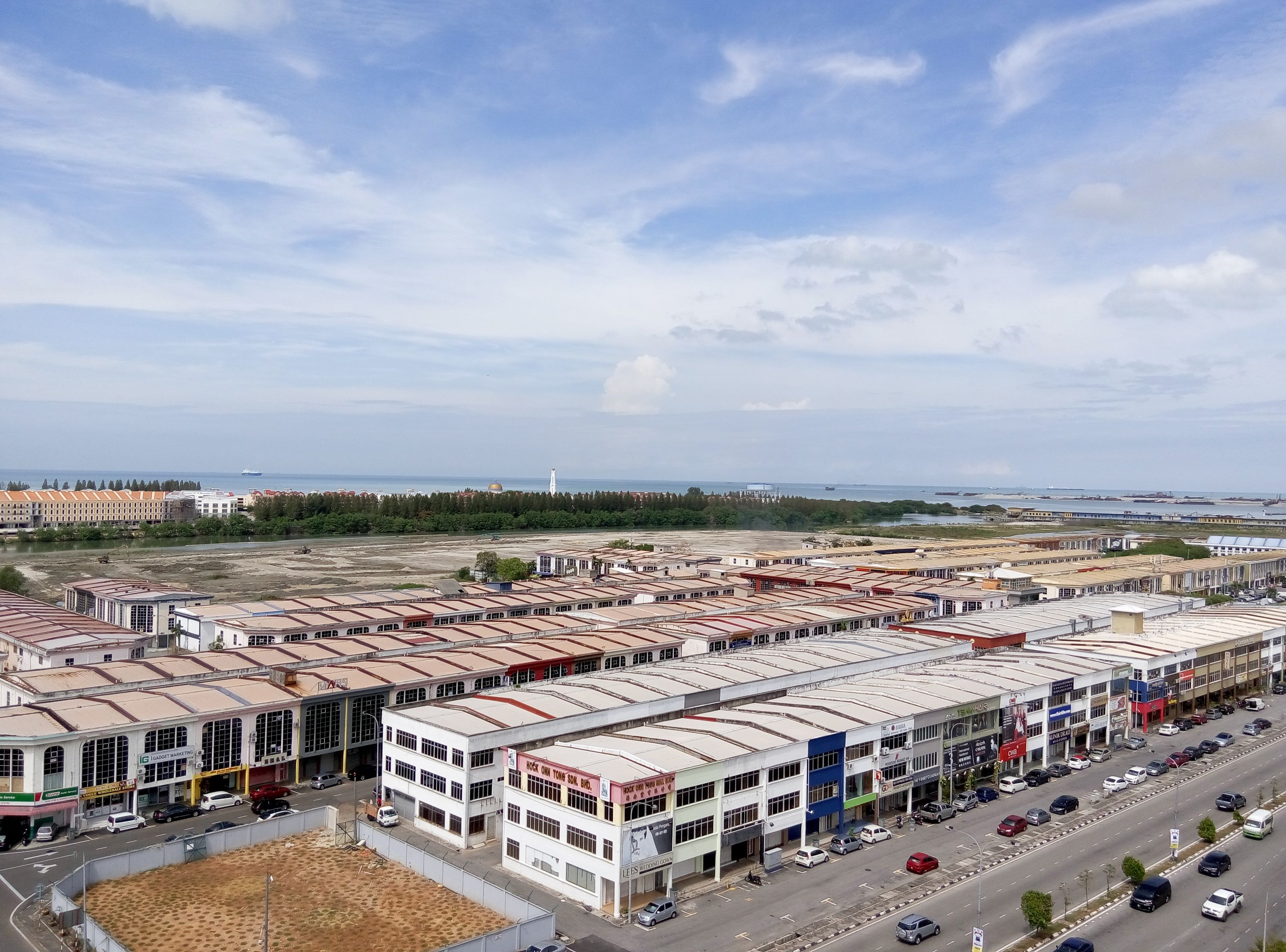 A commercial area with multiple buildings, a road with parked cars, and a distant view of the sea.