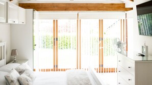 Un dormitorio con una cama grande, vigas de madera en el techo, una araña, un aparador y vistas al exterior con vegetación.