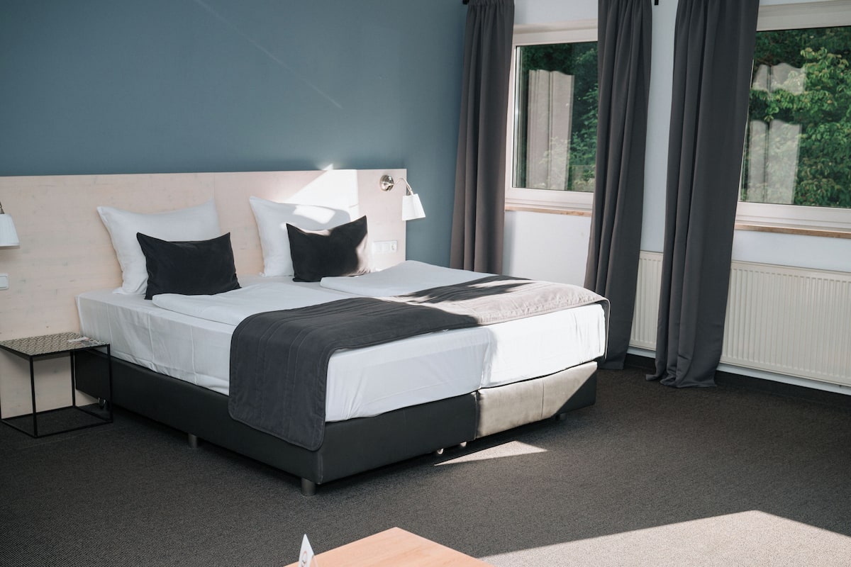 A modern hotel room with a large bed, a bedside table, a lamp, and a window with curtains.