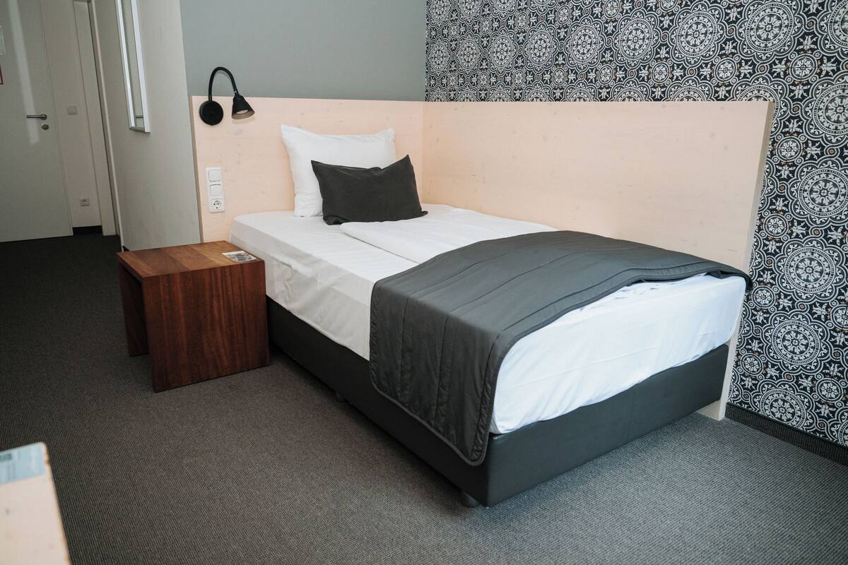 A neatly made bed with a black headboard, a wooden nightstand, a wall-mounted lamp, and patterned wallpaper.