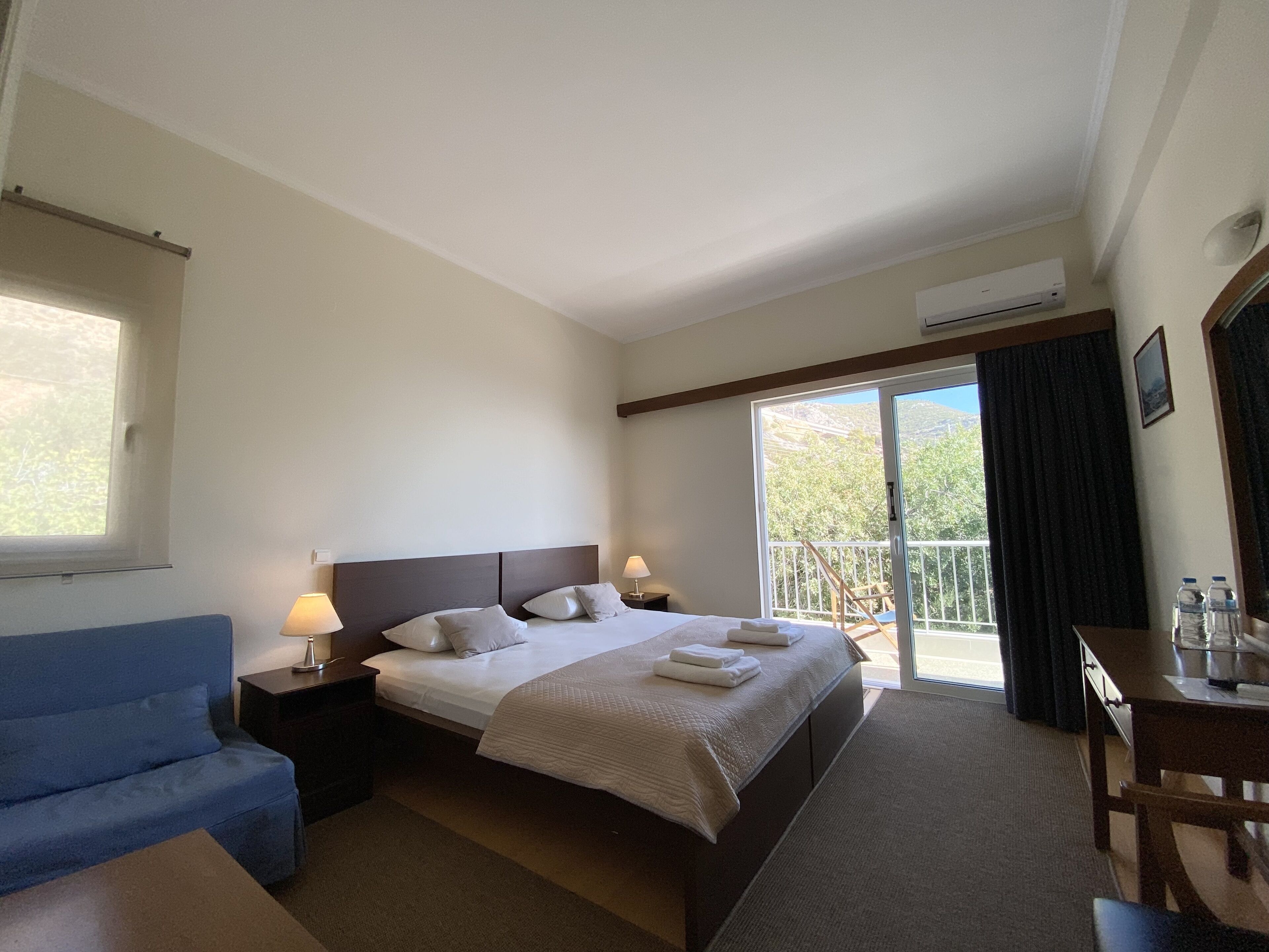 Habitación de hotel con cama, sofá azul, escritorio con silla y balcón con vistas a árboles y montañas.